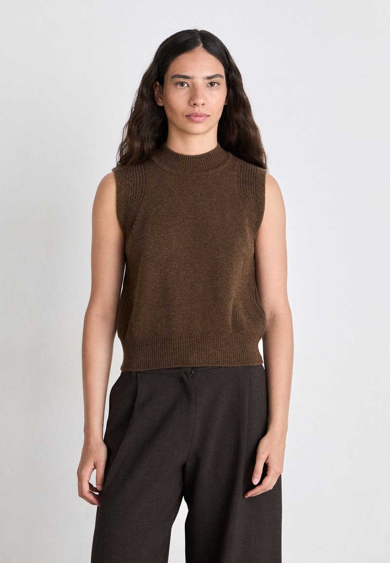 Brauner ärmelloser Strickpullover mit hohem Kragen und geripptem Saum. Getragen mit dunklen, weit geschnittenen Hosen. Einfaches und sauberes Design.