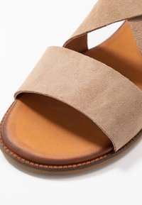 Sandalia de ante beige con dos tiras anchas cruzadas, una plantilla de color bronceado suave y una suela de goma texturada marrón. Diseño minimalista y líneas limpias.