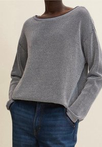 Blauer und grauer, gemusterter Strickpullover mit weitem Ausschnitt, lockerer Passform und langen Ärmeln, kombiniert mit dunkelblauen Jeans.