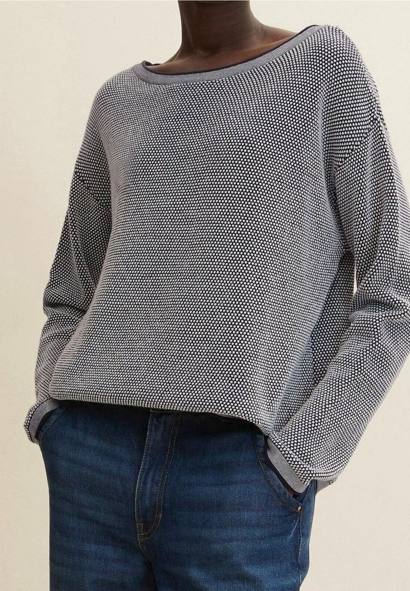 Blauer und grauer, gemusterter Strickpullover mit weitem Ausschnitt, lockerer Passform und langen Ärmeln, kombiniert mit dunkelblauen Jeans.