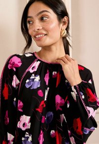 Blusa nera floreale con motivi rosa, rossi e viola. Presenta un colletto arricciato, polsini elasticizzati e rifiniture rosa a contrasto.
