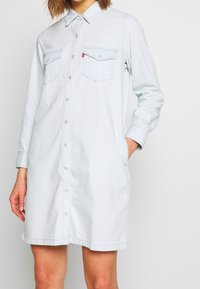 Light blue denim shirt dress with a button front, long sleeves, two chest pockets, and side pockets. Subtle texture, relaxed fit.