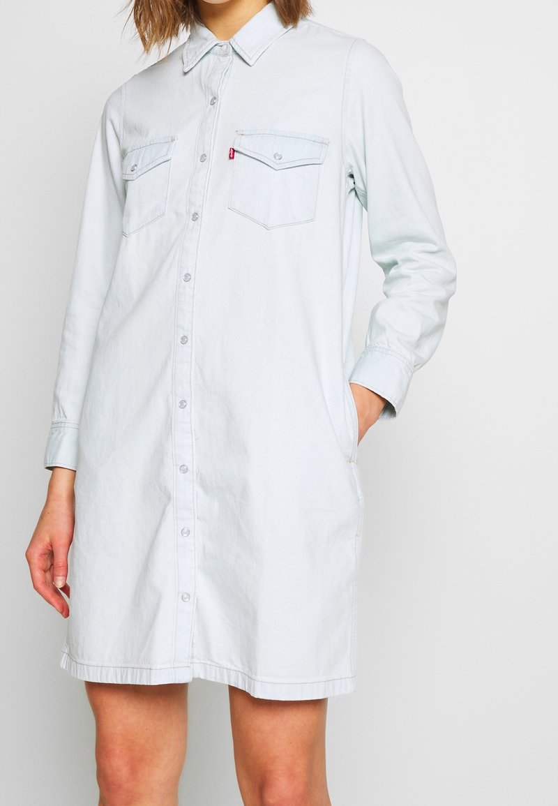 Light blue denim shirt dress with a button front, long sleeves, two chest pockets, and side pockets. Subtle texture, relaxed fit.