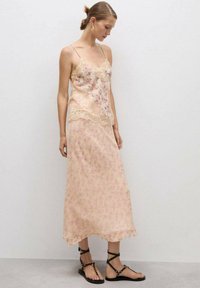 Top floral în piersică deschisă cu dantelă, purtat peste o fustă lungă până la gleznă, având un model delicat de flori. Sandale negre cu curele.