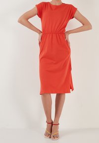 Mujer vestida con un vestido naranja ajustado hasta la rodilla, con mangas cortas y sandalias de tobillo multicolores, de pie frente a un fondo blanco liso.