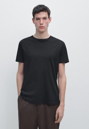 Jonge man met kort donker haar, staand tegen een effen witte muur, gekleed in een zwart T-shirt met korte mouwen en donkerbruine broek.