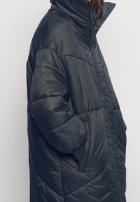 Giacca imbottita in blu scuro, caratterizzata da un colletto alto, chiusura con zip, tasche laterali e un motivo trapuntato per texture e isolamento.