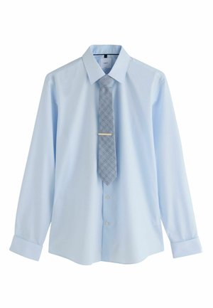 Person fastening silver cufflink on light blue dress shirt with dark trousers visible at waist.