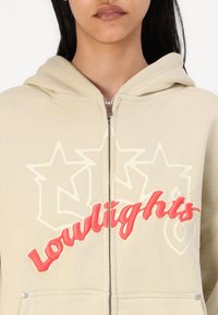 Beige zip-up hoodie featuring a large embroidered graphic in red and cream, with a front pocket and a drawstring hood. Silky texture visible.