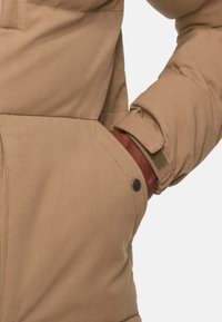 Cappotto beige con una texture di tessuto morbida, dotato di una tasca laterale e un dettaglio con bottoni. I polsini sono regolabili con velcro.