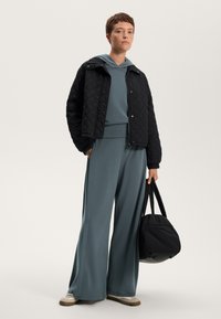 Giacca nera trapuntata sopra una felpa teal e pantaloni larghi abbinati. Il modello tiene una borsa nera. Presenta scarpe con suola in gomma.