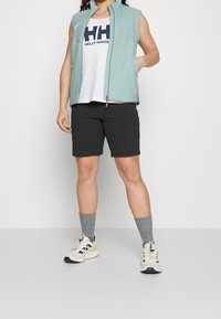 Chaleco sin mangas verde claro sobre una camiseta blanca con texto negro, combinado con pantalones cortos negros y calcetines grises texturizados, completado con zapatillas deportivas.