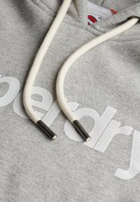 Hoodie gris avec impression en texte craquelé blanc "superdry," doté de cordons crème et de poignées en métal, mettant en valeur une texture douce.