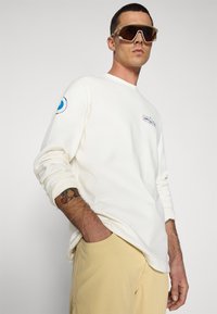 Cream long-sleeve shirt with a colorful logo on the chest and blue accent on the left sleeve, paired with light tan pants and sunglasses.