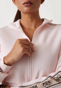 Svijetlopinkasta jakna na zip s visokim ovratnikom, mekog materijala, s "Guess" logotipom i uzorkovanim naglascima duž rukava.