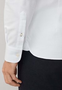 Chemise blanche à manches longues avec un poignet pointu, comportant deux boutons. La texture visible est lisse, le tissu est léger et respirant.