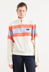 Kremfarget half-zip sweatshirt med oransje og lyseblå horisontale striper. Har en logo på brystet og avslappet passform med ribbestrikkede mansjetter.