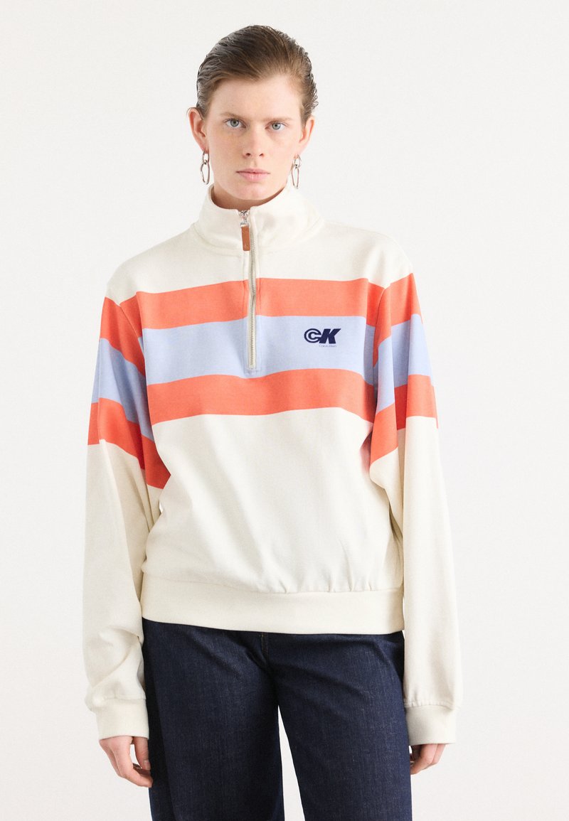 Kremfarget half-zip sweatshirt med oransje og lyseblå horisontale striper. Har en logo på brystet og avslappet passform med ribbestrikkede mansjetter.
