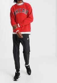 Röd sweatshirt med "NIKE AIR" i stora svarta bokstäver, parat med svarta byxor. Har vita detaljer vid kragen och ärmarna. Svarta skor.