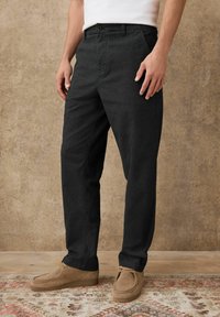 Man wearing black corduroy pants, beige suede lace-up shoes, standing on patterned rug against textured tan wall.