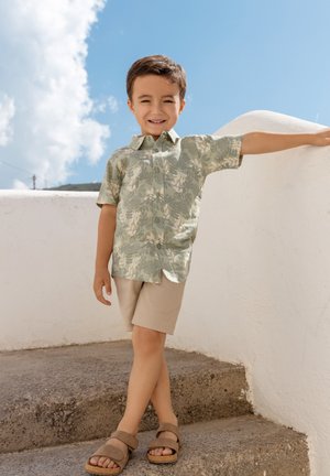 Un jeune garçon portant une chemise à manches courtes à motifs et un short se tient sur des marches extérieures, appuyé contre un mur blanc sous un ciel bleu parsemé de nuages.