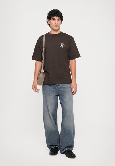 Uomo con capelli ricci che indossa una T-shirt marrone scuro, jeans a gamba larga blu chiaro, scarpe nere e porta una borsa a tracolla beige.