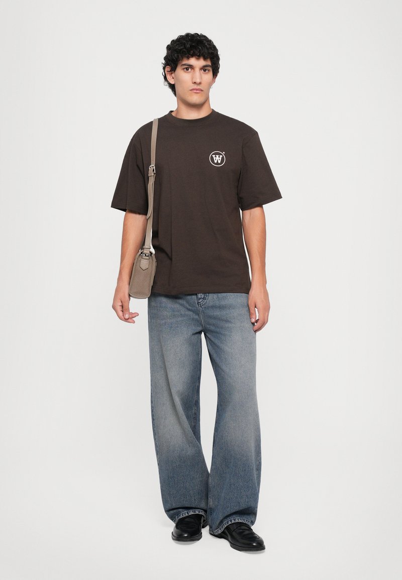 Mann mit lockigen Haaren, der ein dunkelbraunes T-Shirt, hellblaue weit geschnittene Jeans, schwarze Schuhe trägt und eine beigefarbene Umhängetasche trägt.