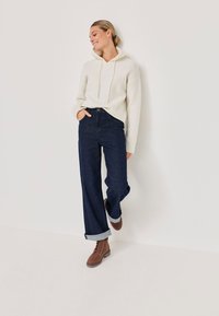 Femme appuyée contre un mur, portant un sweat à capuche blanc, un jean large à ourlet foncé et des bottes brunes à lacets, souriante et regardant en bas.