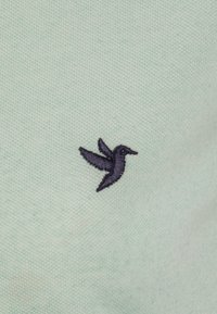 Lichtgroene stof met een textuurafwerking heeft een navyblauwe geborduurde kolibrilogo aan de linkerkant.