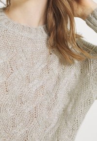 Pull en maille beige clair avec un motif de torsades texturé et une légère brillance. Il présente un col rond et une coupe ample.