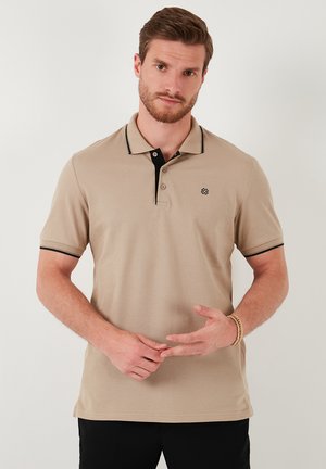 Bež polo majica s crnom kragnom i rubovima, kratkim rukavima, teksturiranim materijalom, otvorom s tri gumba i logotipom na lijevoj strani prsiju.