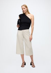 Čierny top na jedno rameno v kombinácii s béžovými širokými nohavicami. Modelka drží čiernu kabelku a nosí čierne sandále na podpätku s remienkami.