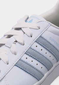 Baskets blanches avec des bandes en daim bleu clair, des perforations, des lacets blancs épais et la marque "SUPERSTAR" sur le côté.