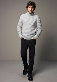 Grauer Strickpullover mit Rollkragen und Diamantmustern, kombiniert mit schwarzen Hosen und schwarzen Schnürschuhen. Das Modell steht mit den Händen in den Taschen.