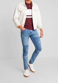 Vit denimjacka över en vinröd pikétröja med vita ränder, kombinerad med slitna blå jeans och vita sneakers med bruna sulor.