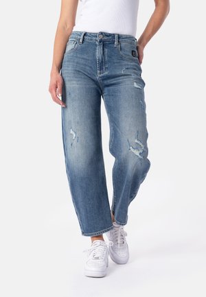 Person trägt hellblaue, abgenutzte Jeans mit geradem Bein, weiße Sneakers und ein in die Hose gestecktes weißes Hemd, steht mit einer Hand auf der Hüfte.