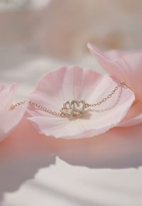 Bracciale d'oro con design a cuore intrecciato in argento, poggiato su petali di fiori rosa, evidenziando texture morbide e colori tenui.