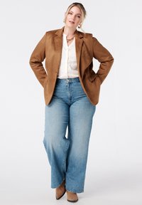 Blazer en daim marron sur une chemise blanche à manches longues, associé à un jean large bleu clair et des escarpins à bouts pointus beiges.