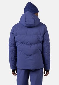 Fioletowa ocieplana kurtka z wysokim kołnierzem i przypiętym kapturem. Posiada pikowane przeszycia, elastyczne mankiety oraz gładką fakturę zapewniającą izolację.