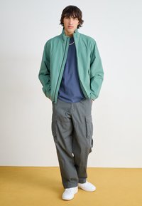 Light green zip-up jacket over a navy blue shirt, paired with gray cargo pants and white sneakers, set against a neutral background.