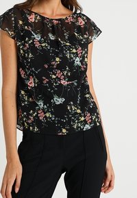 Svart blommig blus med korta, genomskinliga ärmar. Har flerfärgade blommönster, lättviktig tyg och en avslappnad passform. Matchas med svarta byxor.