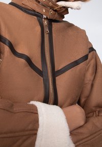 Veste brune avec une texture lisse, présentant des accents en cuir noir et une fermeture éclair à l'avant. Le col est doublé d'une douce polaire blanche.
