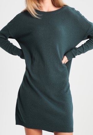 Frau trägt ein dunkelgrünes, langärmeliges Strickkleid mit Knopfdetails an einer Schulter, die Hände auf den Hüften ruhend.