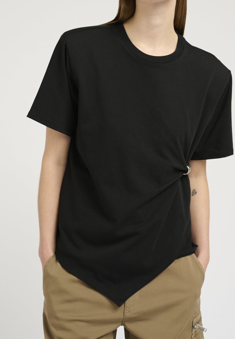 Schwarzes Baumwoll-T-Shirt mit lockerer Passform, das an der Vorderseite einen geknoteten Detail und einen abgerundeten Saum besitzt. Einfache Textur, kurze Ärmel.
