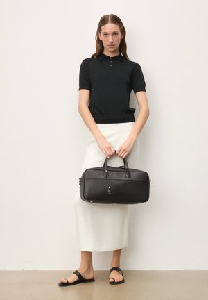 Čierna kožená kabelka s dvoma rúčkami a textúrovaným povrchom, držená modelkou v krátkom rukáve čierneho topu a bielej sukni.