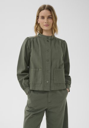 Femme aux cheveux blonds mi-longs portant une veste vert olive boutonnée avec deux poches avant et un pantalon assorti, mains dans les poches.