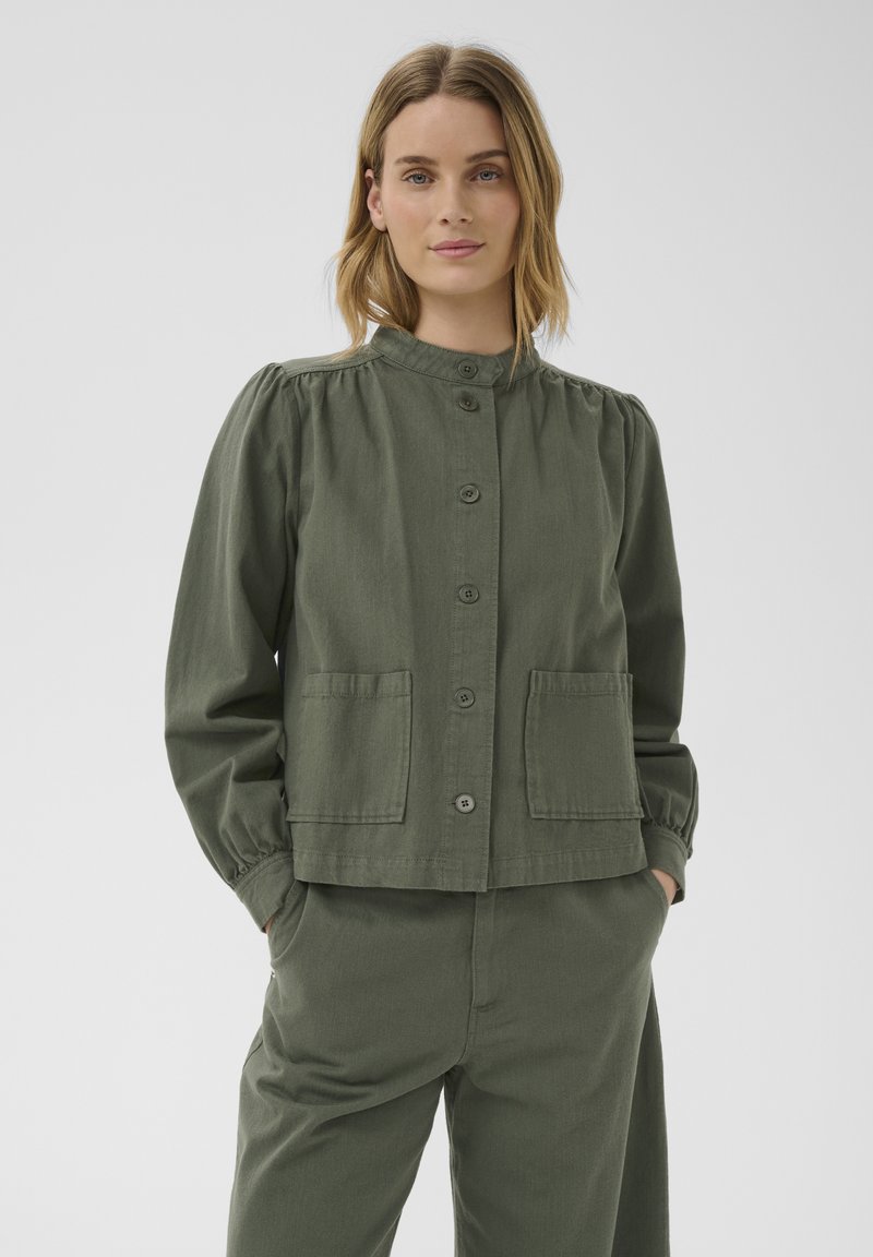 Femme aux cheveux blonds mi-longs portant une veste vert olive boutonnée avec deux poches avant et un pantalon assorti, mains dans les poches.
