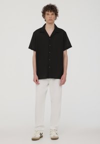 Camicia nera a maniche corte con bottoni e colletto, abbinata a pantaloni bianchi a gamba dritta e sneakers beige con dettagli neri. Design semplice e pulito.