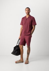 Burgundi spalvos trumpomis rankovėmis marškiniai su tekstūruotu audiniu, derančios šortai ir rudos šlepetės. Laiko juodą krepšį su sidabro detalėmis.