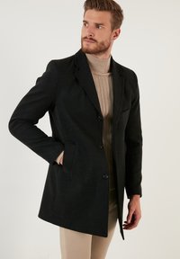 Abrigo de lana negro con un diseño ajustado, botones de un solo pecho, solapas con muesca y bolsillos laterales. Lleva puesto un suéter de cuello alto acanalado en color beige.